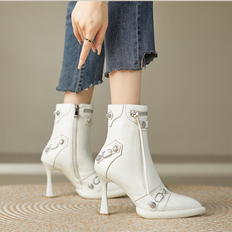 White high-heeled ankle boots with decorative elements worn by a person on a neutral background.